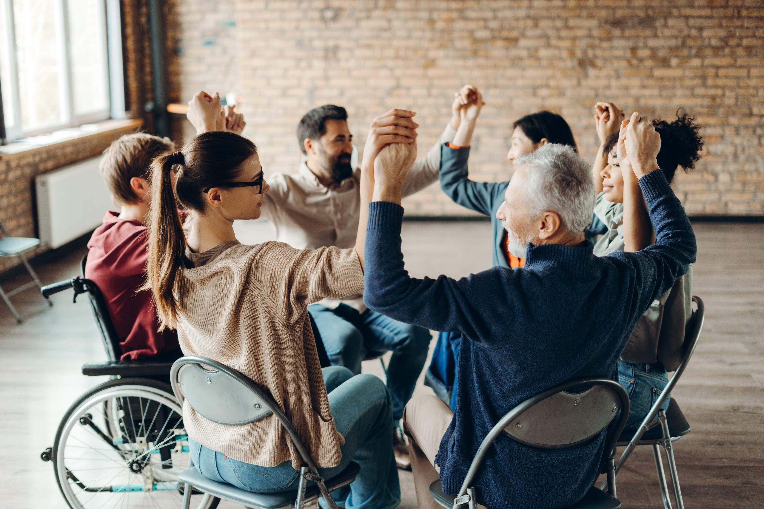 Diverse group of smiling multinational people including person using wheelchair, sitting in circle, holding hands teamwork during meeting. Mental health care concept